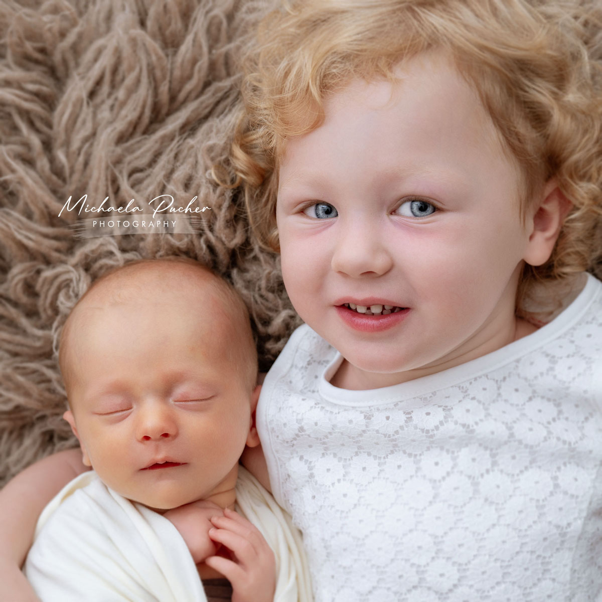 Älteres Geschwisterkind liegt lächelnd neben seinem schlafenden neugeborenen Geschwisterchen auf einer flauschigen Decke.